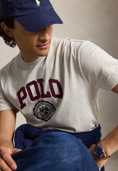 Νέος άνδρας που φοράει γκρι μπλουζάκι Polo Ralph Lauren, μπλε σκουφάκι με λογότυπο, μπλε πουλόβερ δεμένο στη μέση και ρολόι με καφέ δερμάτινο λουράκι.