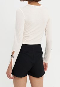 Weißes, geripptes Langarmoberteil mit ausgestellten Ärmelabschlüssen, die Ausschnitte und schwarze Schleifenakzente aufweisen, kombiniert mit hochgeschnittenen schwarzen Shorts.