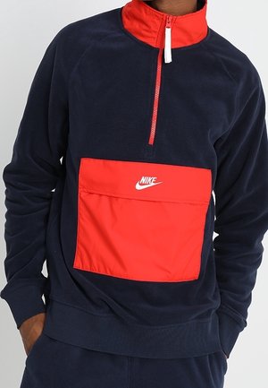 Mężczyzna ubrany w granatowo-czerwony pulower Nike z przednim zamkiem i dużą czerwoną kieszenią z białym logo Nike.