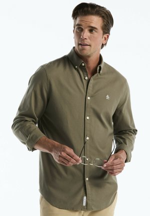 Man wearing olive green button-up shirt holding eyeglasses, looking to the side against a plain light background.