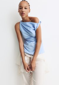 Ung kvinde med fletninger iført en off-shoulder lyseblå top og cremefarvede teksturerede bukser, lænet frem mod en hvid væg.