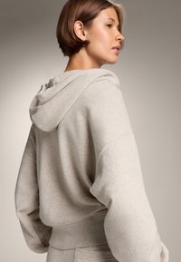 Felpa grigia chiara realizzata in tessuto morbido, con una vestibilità morbida, cappuccio e polsini a coste. Texture liscia con dettagli minimi.