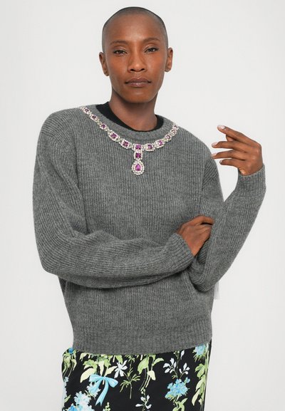 Grauer, gestrickter Pullover mit geripptem Muster, ausgestattet mit einem mit lila und klaren Steinen besetzten Kragen. Das Model posiert mit verschränkten Armen.