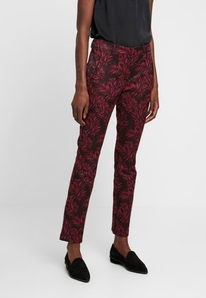 Pantalon bordeaux ajusté à motifs de feuilles rouges porté avec un haut sans manches noir et des mocassins noirs sur une personne debout.