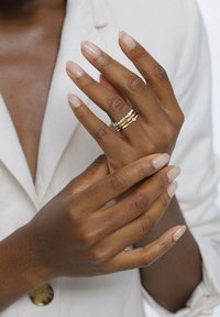 Nahaufnahme von zwei Händen mit manikürten Nägeln, die drei Ringe zeigen – zwei silberne und einen goldenen – vor einer hellen, maßgeschneiderten Bluse.