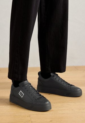 Czarne trampki z fakturowanym wykończeniem, podwyższoną podeszwą i wstążkowymi sznurowadłami. Z boku znajduje się kwadratowy akcent. W zestawieniu z czarnymi spodniami.