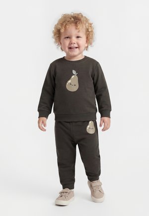 Tout-petit avec des cheveux blonds bouclés portant un sweat-shirt et un pantalon foncés assortis avec des illustrations de poires souriantes, des baskets beiges, debout sur fond blanc.