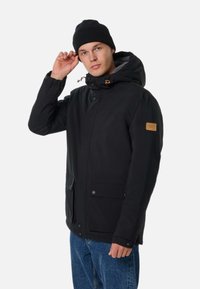 Giovane uomo che indossa una giacca con cappuccio nera e un berretto di lana nero, aggiusta il berretto con una mano, in piedi davanti a uno sfondo bianco uniforme.
