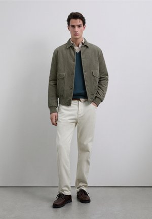 Giacca in velluto a coste verde oliva con bottoni e due tasche anteriori, indossata sopra un maglione scuro, abbinata a pantaloni color crema e scarpe marroni.