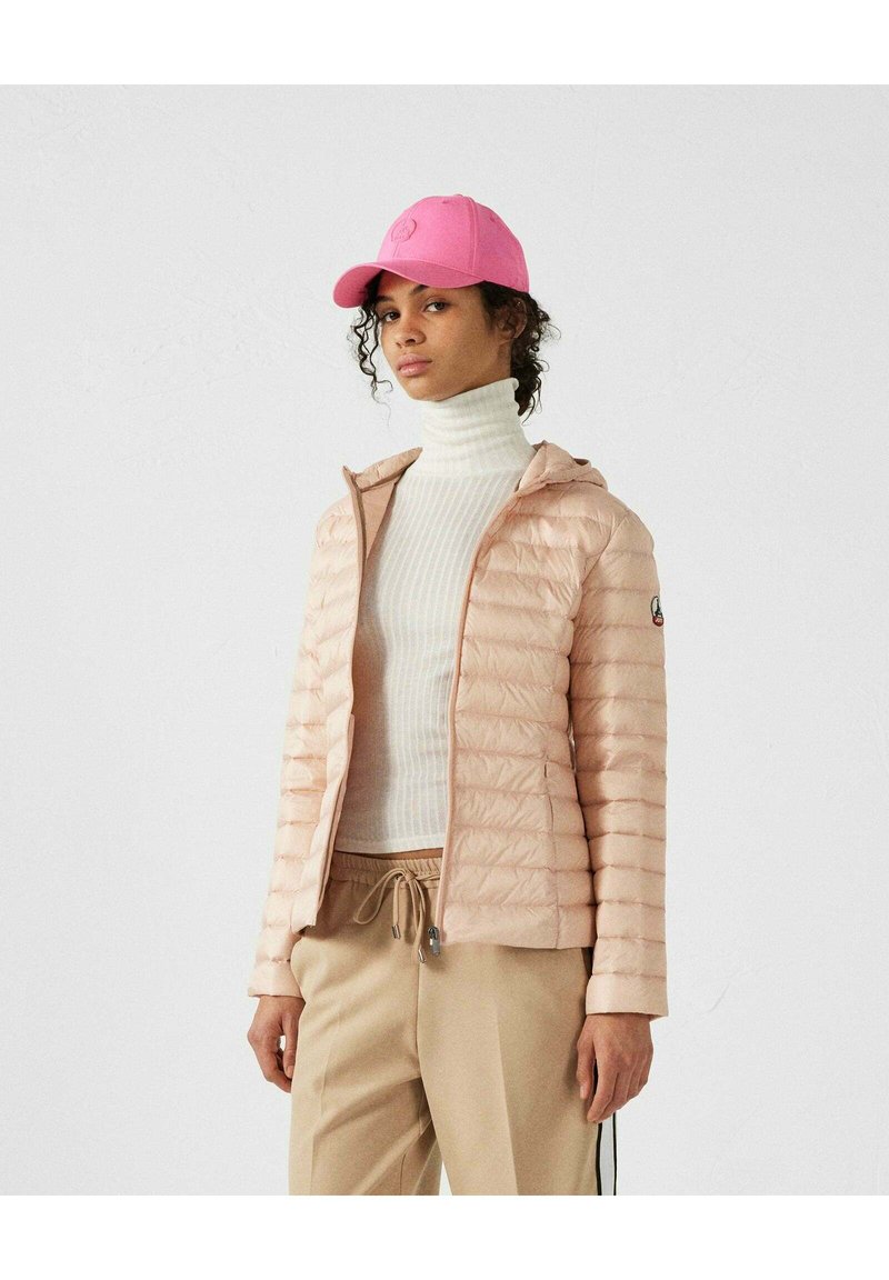 Giacca puffer rosa chiaro con zip frontale e cappuccio, indossata sopra un dolcevita bianco a coste. Abbinata a pantaloni beige con lacci. Berretto rosa.
