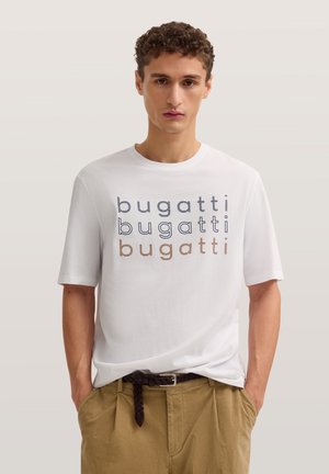Junger Mann trägt ein weißes T-Shirt mit dreimal in verschiedenen Farben bedrucktem „bugatti“, beigefarbene Hose und geflochtenen Gürtel, Hände in den Taschen.
