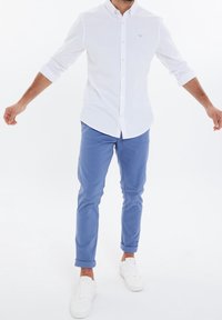 Camicia bianca con bottoni, maniche lunghe e colletto, abbinata a pantaloni tapered azzurri e sneakers bianche. Tessuto morbido e design semplice.