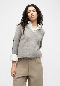 Γυναίκα με σγουρά μαλλιά, φορώντας γκρι πουλόβερ με V λαιμόκοψη πάνω από λευκό πουκάμισο με μανσέτες και μπεζ παντελόνι ψηλής μέσης, στέκεται μπροστά σε απλό φόντο.