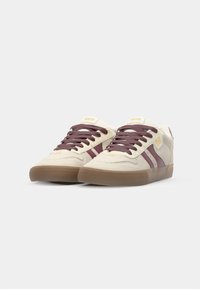 Beige sneakers with maroon stripes and laces, displaying a brown rubber sole. The design features a branded logo on the side.