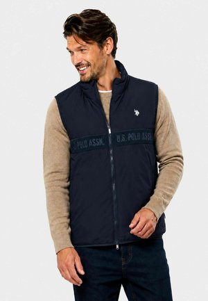 Mand der smiler, iført navyblå U.S. Polo Assn. vest over beige sweater og mørke jeans, står mod en ensartet lys baggrund.