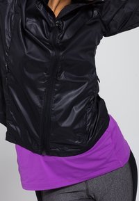 Czarna, lekka, zapinana kurtka o gładkiej fakturze, noszona na purpurowej siatkowej bluzce. Kurtka ma boczne kieszenie i dopasowany krój.