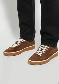 Brown suede low-top sneakers with white laces, rubber sole, and contrasting beige midsole. Notable stitching and a smooth texture.