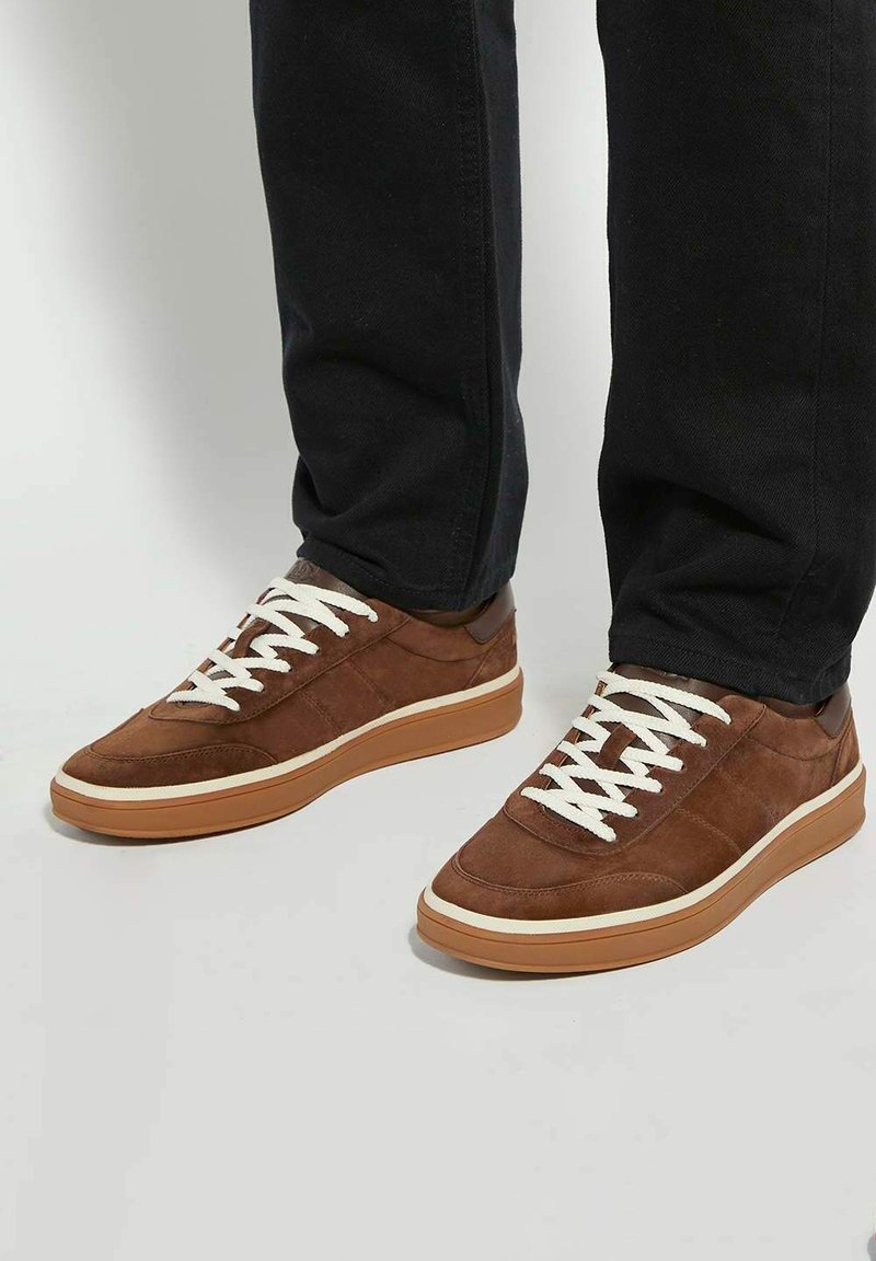 Brown suede low-top sneakers with white laces, rubber sole, and contrasting beige midsole. Notable stitching and a smooth texture.