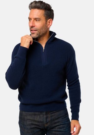 Mann in marinblauer Zip-Up-Jacke und dunklen Jeans, der den Reißverschlussgriff nahe am Kinn hält und zur Seite blickt, vor einem schlichten Hintergrund.