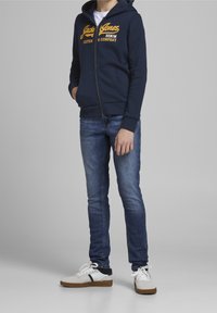 Marinefarbener Zip-Hoodie mit gelbem Logo, dunkle Jeans und weiße Schuhe mit grauen Akzenten und schwarz gestreiftem Design. Leger, enganliegendes Design.