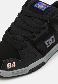 Sapatilha preta feita de malha e material sintético. Apresenta detalhes cinzentos, logo na lateral e "94" impresso em vermelho na ponta.