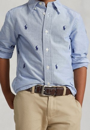 Chłopiec w jasnoniebieskiej koszuli na guziki z granatowymi logo gracza polo, podwiniętymi rękawami, beżowych spodniach oraz brązowym pasku z srebrną klamrą.