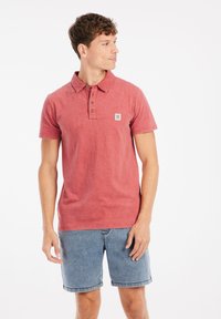 Rode polo-shirt van textiel met een gestructureerde stof, met een drieknoopsluiting en een klein logo-embleem, gecombineerd met lichtblauwe denimshorts.