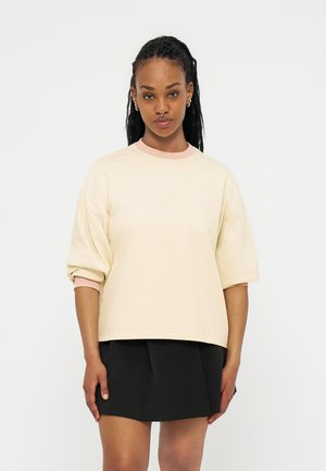 Junge Frau mit langem, geflochtenem Haar, die einen cremefarbenen Pullover mit rosa Bündchen und Kragen sowie einen kurzen schwarzen Rock trägt, vor weißem Hintergrund stehend.