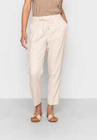 Pantalon beige léger avec taille élastique et cordon, équipé de deux poches latérales et de jambes fuselées. Porté avec des sandales plates à bout ouvert.