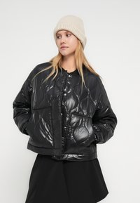 Νεαρή γυναίκα που φοράει εκρού πλεκτό σκουφάκι, μαύρο γυαλιστερό μπουφάν με σχέδιο καπιτονέ και κουμπιά με τρούκ, και μαύρη φούστα, κοιτάζει προς τα αριστερά της.