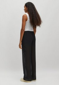Zwarte geplooide broek met een relaxte pasvorm en een elastische tailleband, gecombineerd met een witte mouwloze top. Het model heeft lang krullend haar.
