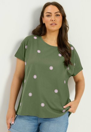Kvinde med langt mørkt hår iført grøn T-shirt med lyserøde tusindfryd og lyseblå jeans, står op ad en lys væg.