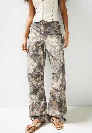 Pantalons à motif camouflage dans des tons terreux atténués, avec une coupe décontractée, une taille à cordon de serrage et des poches latérales. Portés avec des tongs.