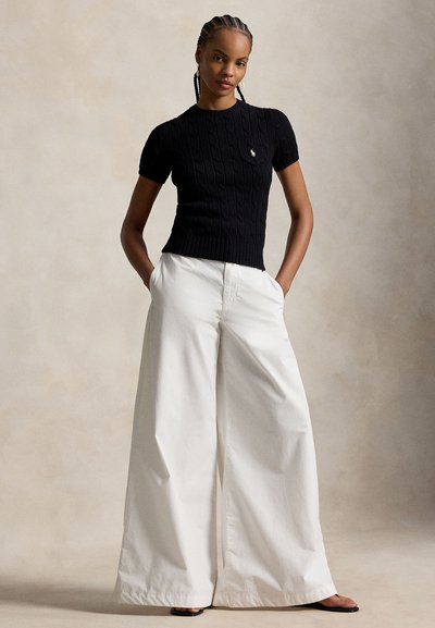 Mujer con cabello trenzado que lleva una camiseta negra de punto de manga corta y pantalones blancos de pierna ancha, de pie con las manos en los bolsillos frente a un fondo liso.