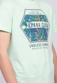 T-shirt en coton vert clair avec un graphisme hexagonal représentant des feuilles tropicales et le texte : "HAVANE CUBA" et "ÉTÉ SANS FIN" en bleu marine.