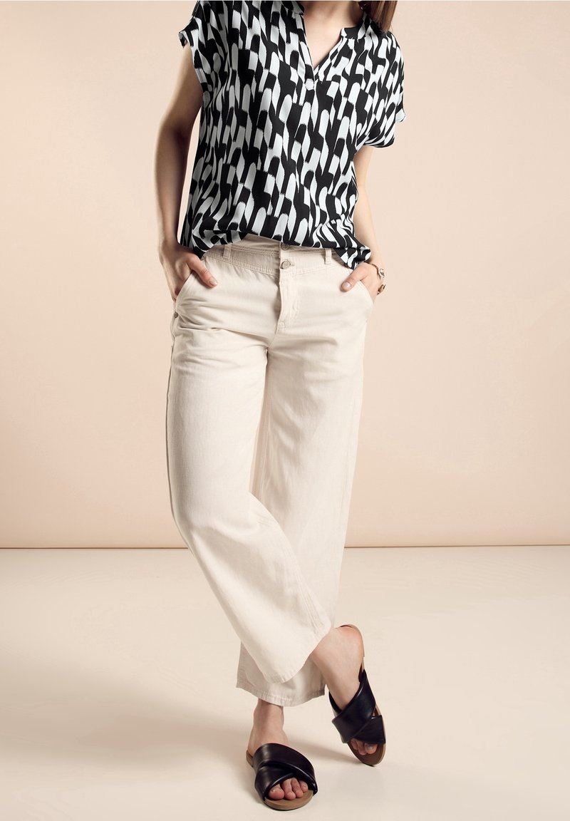 Camicia a maniche corte con motivo in bianco e nero, abbinata a pantaloni larghi beige chiaro e sandali neri a infradito. Sfondo chiaro.