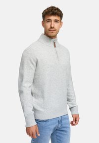 Ljusgrå tröja med hög krage och halvdragkedja, tillverkad av ett mjukt, texturerat tyg. Har ribbade manschetter och nederkant. Bärs med blå jeans.