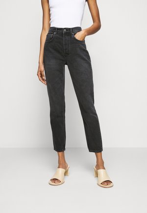 Kobieta nosząca wysokie czarne dżinsy zwężane ku dołowi, biały top bez rękawów oraz beżowe sandały na obcasie typu bloczek z odkrytymi palcami, stoi z jedną ręką w kieszeni.