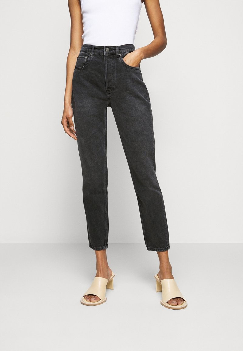 Femme portant un jean noir taille haute avec jambes fuselées, un haut blanc sans manches, et des sandales à talons blocs beige à bout ouvert, debout avec une main dans la poche.