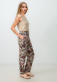 Débardeur beige, pantalon ample à motifs floraux en rouge, violet et vert. Le mannequin se tient avec une main dans une poche.