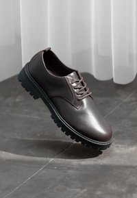 Dark brown leather lace-up shoe with thick black sole on gray tiled floor in front of sheer white curtains.