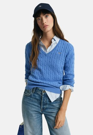Blue cable-knit sweater with a V-neck, worn over a light blue striped shirt, paired with faded blue jeans. Accented with a navy cap.