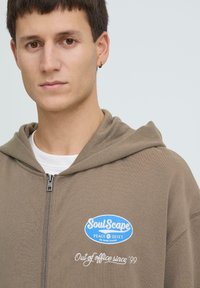 Sweat à capuche marron zippé avec un patch logo bleu indiquant "SoulScape." Présente une broderie blanche indiquant "Out of office since '99." Texture douce.