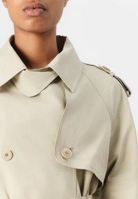 Beige takki, jossa on leveä kaulus, kaksiriviset napit ja olkatoppaukset. Sujuva kangas, jossa näkyvät ompeluyksityiskohdat.