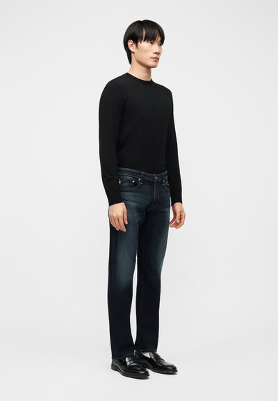 Νεαρός άνδρας όρθιος, φορώντας μαύρο πουκάμισο με μακριά μανίκια, σκούρο μπλε τζιν και μαύρα δερμάτινα παπούτσια, μπροστά σε μια απλή ανοιχτόχρωμη επιφάνεια.