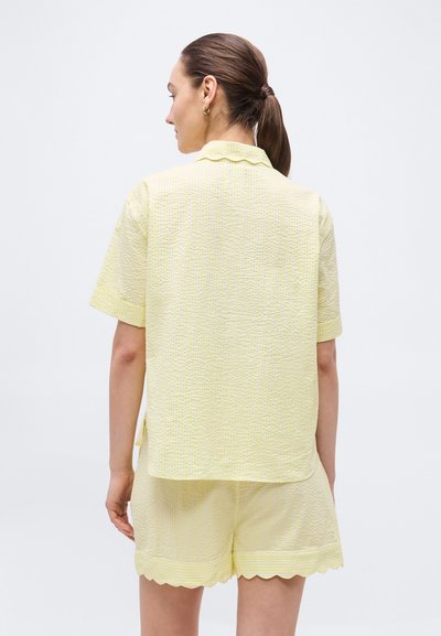 Γυναίκα με καστανή αλογοουρά που φοράει ανοιχτόκίτρινο υφασμάτινο κοντομάνικο τοπ και αντίστοιχα σορτς με κυματιστά τελειώματα, γυρισμένη προς τα πίσω πάνω σε απλό φόντο.