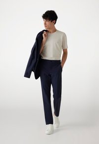 Marineblaue Hose, hellbeige T-Shirt und weiße Sneakers. Das Model trägt einen marineblauen Blazer und hat ein dezentes Logo auf dem T-Shirt.