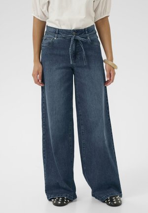 Femeie purtând blugi evazați de culoare închisă cu centură, bluză albă cu mâneci bufante, brățară groasă și pantofi negri cu ținte.