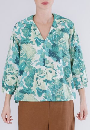 Kvinde iført en løs, blomsterprintet bluse i nuancer af grøn og teal med elastiske trekvartærmer og brune bukser.