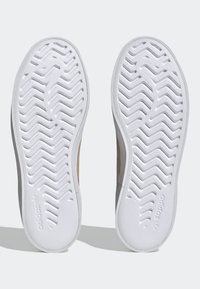 Valkoiset tennarit, joissa on sahalaitakuvioiset pohjat ja Adidas-logo näkyvissä kunkin pohjan kantapääalueella, esitettynä alapuolelta katsottuna.